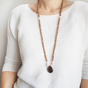 A woman stands in a white sweater wearing a sandalwood mala necklace. The mala has a teardrop shaped Smoky Quartz  guru bead. Above the guru bead are rose quartz and light pink moonstone beads followed by 6mm sandalwood beads separated by 2mm silver beads.
