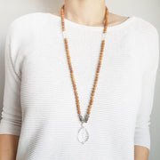 A woman stands in a white sweater wearing a sandalwood mala necklace. The mala has a faceted teardrop shaped Clear Quartz guru stone . Above the guru stone is a small mother of pearl bead and four roundel shaped labradorite beads going up on each side. The Labradorite has flashes of blue and green. The rest of the mala necklace is made with 6mm sandalwood beads separated by 2mm silver spacer beads.  