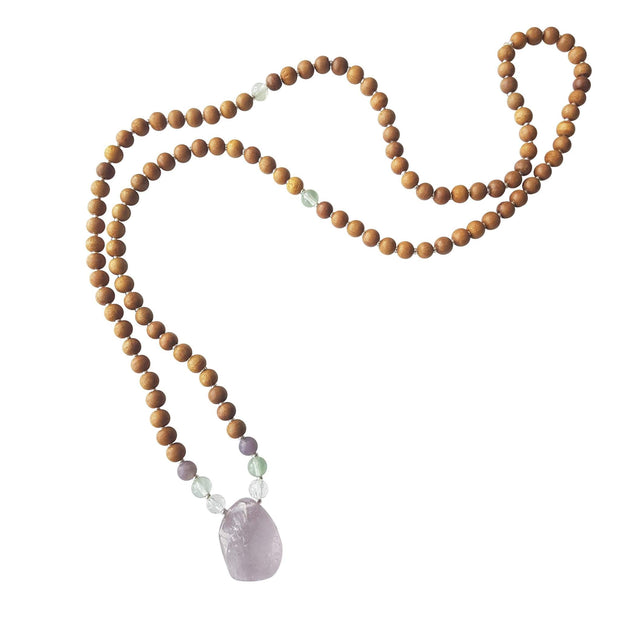 Close up image on a white background of a mala necklace. The mala has a slightly squared organic shaped light purple Amethyst Guru Bead . On each side above the guru stone, is one clear quartz, one green fluorite and one purple lepidolite bead. The rest of the mala is made with 6mm sandalwood beads separated by 2mm silver spacer beads. In the middle of the necklace there is a single green fluorite bead on each side.