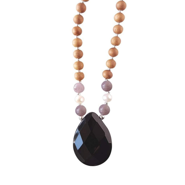Close up image on a white background of a mala bead necklace. The mala has a pear shaped faceted onyx guru bead.   On each side above the Onyx guru is one purple lepidolite bead, one pearl and one more lepidolite bead. The rest of the mala is made with 6mm sandalwood beads separated by 2mm silver spacer beads.
