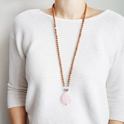 A woman stands in a white sweater wearing a sandalwood mala necklace. The mala has a teardrop shaped Rose Quartz guru bead. Above the guru bead are lepidolite, dalmation jasper and clear quartz beads followed by 6mm sandalwood beads separated by 2mm silver beads.