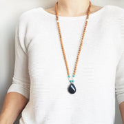 A woman stands in a white sweater wearing a sandalwood mala necklace. The mala has a teardrop shaped Black Onyx guru bead . Above the guru bead are pearl and turquoise jasper beads followed by 6mm sandalwood beads separated by 2mm silver beads.