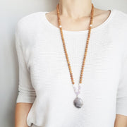 A woman stands in a white sweater wearing a sandalwood mala necklace. The mala has an oval shaped Lepidolite guru bead . Above the guru bead are moonstone, amethyst and rainbow moonstone beads followed by 6mm sandalwood beads separated by 2mm silver beads.