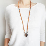 A woman stands in a white sweater wearing a sandalwood mala necklace. The mala has a pear shaped Smoky Quartz guru bead . Above the guru bead are pink moonstone and labradorite beads followed by 6mm sandalwood beads separated by 2mm silver beads.