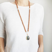 A woman stands in a white sweater wearing a sandalwood mala necklace. The mala has a large 22x30mm teardrop shaped Labradorite guru bead with a border of gold.  The Labradorite has flashes of green and gold . Above the guru bead are Pink Moonstone, Rainbow Moonstone and Aquamarine beads followed by 6mm sandalwood beads separated by 2mm gold beads.