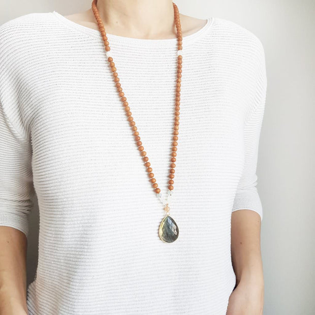 A woman stands in a white sweater wearing a sandalwood mala necklace. The mala has a large 22x30mm teardrop shaped Labradorite guru bead with a border of gold.  The Labradorite has flashes of green and gold . Above the guru bead are Pink Moonstone, Rainbow Moonstone and Aquamarine beads followed by 6mm sandalwood beads separated by 2mm gold beads.