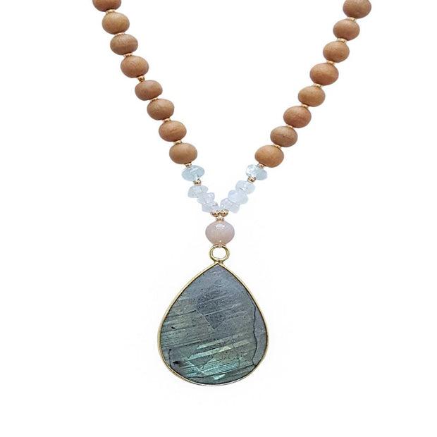 Close up image on a white background of a mala necklace with a large 22x30mm teardrop shaped Labradorite guru bead with a border of gold.  The Labradorite has flashes of green and blue . Above the guru bead are Pink Moonstone, Rainbow Moonstone and Aquamarine beads followed by 6mm sandalwood beads separated by 2mm gold beads.