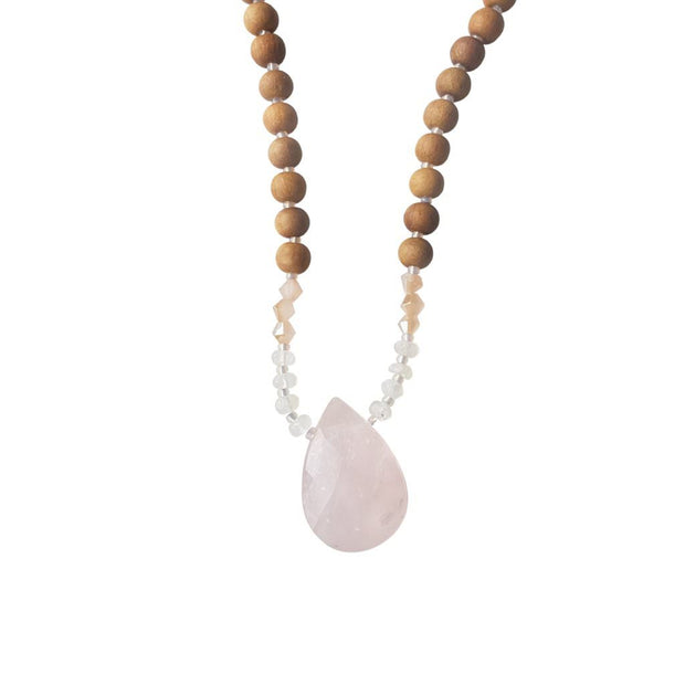 Close up image on a white background of a mala necklace with a faceted teardrop shaped Rose Quartz guru stone . Above the guru stone are small moonstone roundel beads and diamond shaped peach coloured beads followed by 6mm sandalwood beads separated by 2mm iridescent pink glass spacer beads. 