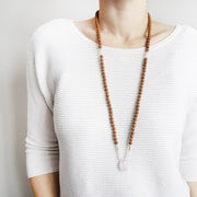 A woman stands in a white sweater wearing a sandalwood mala necklace. The mala has a faceted teardrop shaped Rose Quartz guru stone . Above the guru stone are small moonstone roundel beads and diamond shaped peach coloured beads followed by 6mm sandalwood beads separated by 2mm iridescent pink glass spacer beads. 