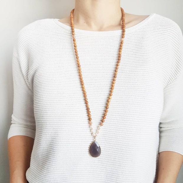 A woman stands in a white sweater wearing a sandalwood mala necklace. The mala has a faceted teardrop shaped Grey Moonstone guru stone edged with gold . On each side above the guru stone are three pearl beads and one pink moonstone bead. The rest of the mala necklace is made with 6mm sandalwood beads separated by 2mm gold spacer beads. 