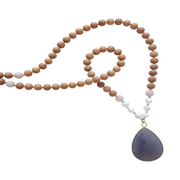 Close up image on a white background of a mala necklace with a 22x30mm faceted teardrop shaped Grey Moonstone guru stone edged with gold . On each side above the guru stone are three pearl beads and one pink moonstone bead. The rest of the mala necklace is made with 6mm sandalwood beads separated by 2mm gold spacer beads. Halfway up the necklace two pink moonstone bead divides the sandalwood into two sections of 27 and one section of 54.