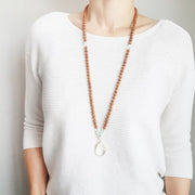A woman stands in a white sweater wearing a sandalwood mala necklace. The mala has a 22x30mm faceted teardrop shaped Clear Quartz guru stone edged with gold . Above the guru stone is one 6mm Rose Quartz bead. On each side above the Rose Quartz are three small roundel aquamarine beads and one small faceted roundel Clear Quartz bead. The rest of the mala is made with 6mm sandalwood beads separated by 2mm gold spacer beads. 