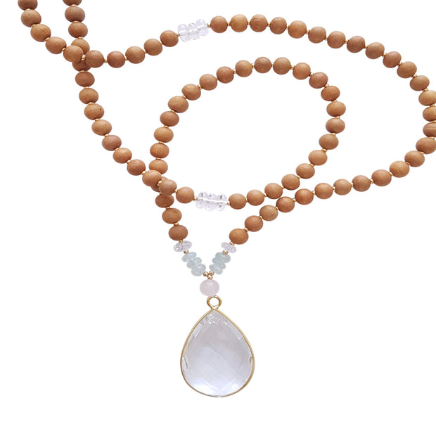 Close up image on a white background of a mala necklace. The mala has a 22x30mm faceted teardrop shaped Clear Quartz guru stone edged with gold . Above the guru stone is one 6mm Rose Quartz bead. On each side above the Rose Quartz are three small roundel aquamarine beads and one small faceted roundel Clear Quartz bead. The rest of the mala is made with 6mm sandalwood beads separated by 2mm gold spacer beads. Halfway up the necklace on each side is a small section of 3 roundel clear quartz beads.