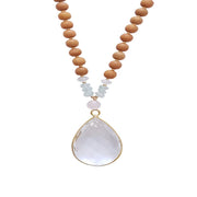 Close up image on a white background of a mala necklace. The mala has a 22x30mm faceted teardrop shaped Clear Quartz guru stone edged with gold . Above the guru stone is one 6mm Rose Quartz bead. On each side above the Rose Quartz are three small roundel aquamarine beads and one small faceted roundel Clear Quartz bead. The rest of the mala is made with 6mm sandalwood beads separated by 2mm gold spacer beads. 