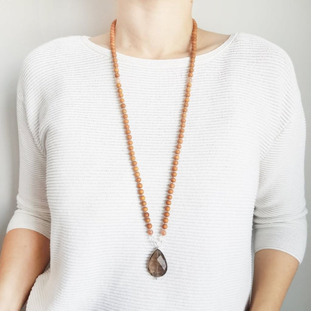 A woman stands in a white sweater wearing a sandalwood mala necklace. The mala has a faceted teardrop shaped Smoky Quartz Guru Bead . On each side above the Guru stone are two round faceted Clear Quartz beads and one round Pink Moonstone bead. The rest of the mala is made with 6mm sandalwood beads separated by 2mm silver spacer beads. 
