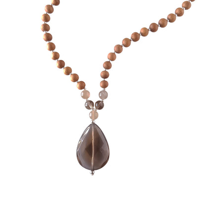 Close up image on a white background of a mala necklace. The mala has a  faceted teardrop shaped Smoky Quartz Guru Bead . Above the Guru stone is a single round grey moonstone bead. On each side, above the grey moonstone, is a 6mm round smoky quartz bead and another grey moonstone bead. The rest of the mala is made with 6mm sandalwood beads separated by 2mm silver spacer beads. 