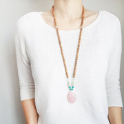 A woman stands in a white sweater wearing a sandalwood mala necklace. The mala has a large faceted teardrop shaped Rose Quartz Guru Bead . On each side above the guru stone, is two 6mm round turquoise beads, 2 serpentine beads and 2 green fluorite beads. The rest of the mala is made with 6mm sandalwood beads separated by 2mm silver spacer beads. In the middle of the necklace there is a single green fluorite bead on each side.