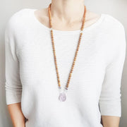 A woman stands in a white sweater wearing a sandalwood mala necklace. The mala has a slightly squared organic shaped light purple Amethyst Guru Bead . On each side above the guru stone, is one clear quartz, one green fluorite and one purple lepidolite bead. The rest of the mala is made with 6mm sandalwood beads separated by 2mm silver spacer beads.  In the middle of the necklace there is a single green fluorite bead on each side.