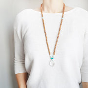 A woman stands in a white sweater wearing a sandalwood mala necklace. The mala has a faceted pear shaped Clear Quartz Guru Bead . On each side above the guru stone, is one turquoise, one light teal amazonite and one rainbow moonstone bead. The rest of the mala is made with 6mm sandalwood beads separated by 2mm silver spacer beads. 