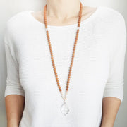 A woman stands in a white sweater wearing a sandalwood mala necklace. The mala has a  faceted pear shaped Clear Quartz Guru Bead . Above the guru stone is a single rainbow moonstone bead. On each side above the rainbow moonstone, is one pink pearl, one white pearl, one diamond shaped peach glass bead and one more pink pearl. The rest of the mala is made with 6mm sandalwood beads separated by 2mm silver spacer beads. In the middle of the sandalwood beads is 1 single clear quartz bead on each side.