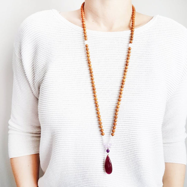 A woman stands in a white sweater wearing a sandalwood mala bead necklace. The mala has a  long gold filled teardrop shaped  Ruby gemstone pendant.  The ruby is a deep burgundy colour and opaque.  Above the ruby is a delicate dark purple lepidolite bead.  On each side above the Lepidolite, are two dainty roundel light purple amethyst beads and three dainty rainbow moonstone beads. The rest of the mala is made with 6mm sandalwood beads separated by 2mm gold spacer beads. 