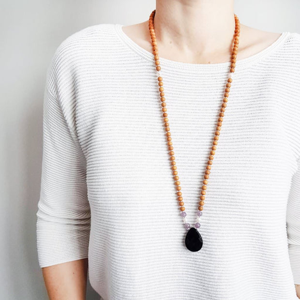 A woman stands in a white sweater wearing a sandalwood mala bead necklace. The mala has a  pear shaped faceted onyx guru bead.   On each side above the Onyx guru is one purple lepidolite bead, one pearl and one more lepidolite bead. The rest of the mala is made with 6mm sandalwood beads separated by 2mm silver spacer beads. 