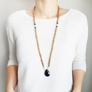 A woman stands in a white sweater wearing a sandalwood mala bead necklace. The mala has a  pear shaped faceted onyx guru bead.   On each side above the Onyx guru is one rose quartz bead and one light teal amazonite bead. The rest of the mala is made with 6mm sandalwood beads separated by 2mm silver spacer beads.  Two thirds of the way up the necklace a amazonite bead flanked by 2 onyx beads break up the sandalwood on each side.