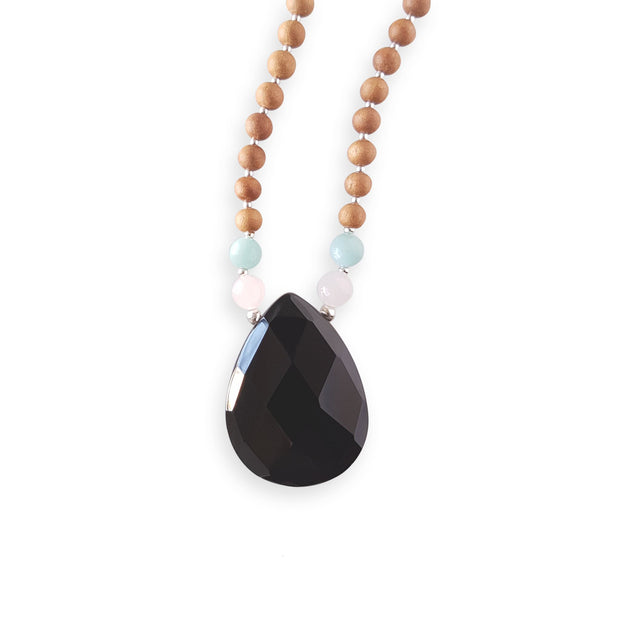 Close up image on a white background of a mala bead necklace. The mala has a pear shaped faceted onyx guru bead.   On each side above the Onyx guru is one rose quartz bead and one light teal amazonite bead. The rest of the mala is made with 6mm sandalwood beads separated by 2mm silver spacer beads. 