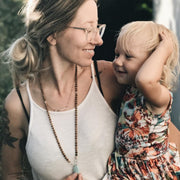 A woman with dark blonde hair and clear rimmed glasses holds her young daughter who has short pale blonde hair and is wearing a floral dress. The woman is smiling at her daughter and holding onto the Amazonite guru bead of the  mala bead necklace she is wearing around her neck. Her daughter is looking at her and smiling. 