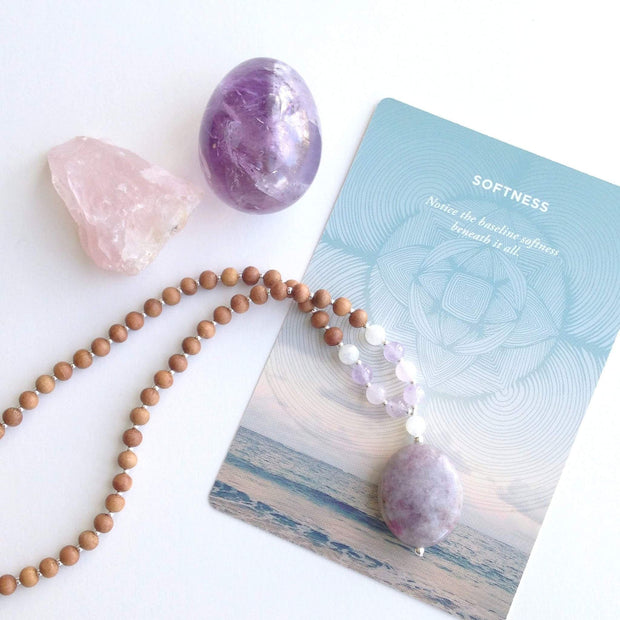 A sandalwood mala necklace is laid on a table over a card that shows a beach with the word softness. A round amethyst crystal and rough cut rose quartz crystal rest beside it. . The mala has an oval shaped Lepidolite guru bead . Above the guru bead are moonstone, amethyst and rainbow moonstone beads followed by 6mm sandalwood beads separated by 2mm silver beads.