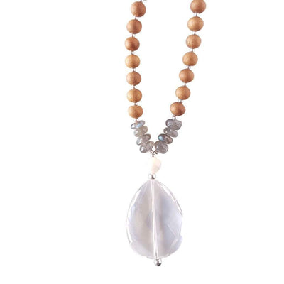 Close up image on a white background of a mala necklace with a faceted teardrop shaped Clear Quartz guru stone . Above the guru stone is a small mother of pearl bead and four roundel shaped labradorite beads going up on each side. The labradorite has flashes of brilliant light blue. The rest of the mala necklace is made with 6mm sandalwood beads separated by 2mm silver spacer beads. 