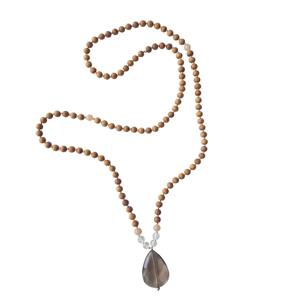 Close up image on a white background of a mala necklace. The mala has a  faceted teardrop shaped Smoky Quartz Guru Bead . On each side above the Guru stone are two round faceted Clear Quartz beads and one round Pink Moonstone bead. The rest of the mala is made with 6mm sandalwood beads separated by 2mm silver spacer beads.  Halfway up the necklace is a single pink moonstone bead on each side.