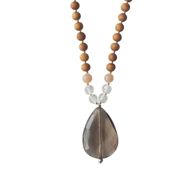 Close up image on a white background of a mala necklace. The mala has a  faceted teardrop shaped Smoky Quartz Guru Bead . On each side above the Guru stone are two round faceted Clear Quartz beads and one round Pink Moonstone bead. The rest of the mala is made with 6mm sandalwood beads separated by 2mm silver spacer beads. 