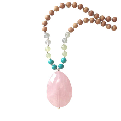 Close up image on a white background of a mala necklace. The mala has a  large faceted teardrop shaped Rose Quartz Guru Bead . On each side above the guru stone, is two 6mm round turquoise beads, 2 serpentine beads and 2 green fluorite beads. The rest of the mala is made with 6mm sandalwood beads separated by 2mm silver spacer beads.