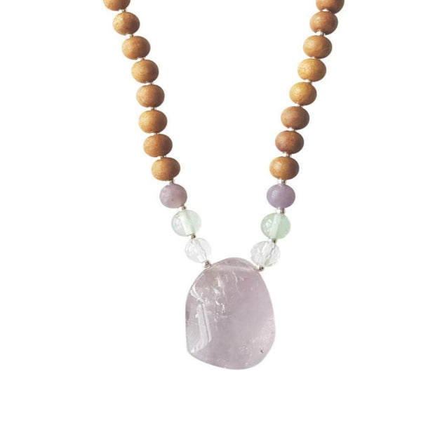 Close up image on a white background of a mala necklace. The mala has a slightly squared organic shaped light purple Amethyst Guru Bead . On each side above the guru stone, is one clear quartz, one green fluorite and one purple lepidolite bead. The rest of the mala is made with 6mm sandalwood beads separated by 2mm silver spacer beads.  