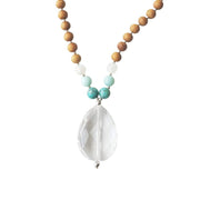 Close up image on a white background of a mala necklace. The mala has a faceted pear shaped Clear Quartz Guru Bead . On each side above the guru stone, is one turquoise, one light teal amazonite and one rainbow moonstone bead. The rest of the mala is made with 6mm sandalwood beads separated by 2mm silver spacer beads. 
