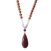 Close up image on a white background of a mala bead necklace. The mala has a long gold filled teardrop shaped  Ruby gemstone pendant.  The ruby is a deep burgundy colour and opaque.  Above the ruby is a delicate dark purple lepidolite bead.  On each side above the Lepidolite, are two dainty roundel light purple amethyst beads and three dainty rainbow moonstone beads. The moonstone has flashes of blue.The rest of the mala is made with 6mm sandalwood beads separated by 2mm gold spacer beads. 